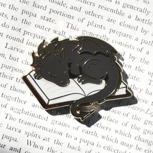 Könnte beinhalten: Schwarze Drachenbrosche mit goldenen Akzenten, die friedlich auf einem weißen Buch schläft. Die Brosche hat detaillierte Flügel und eine glatte, polierte Oberfläche. Ein charmantes Accessoire für Fantasy-Enthusiasten.