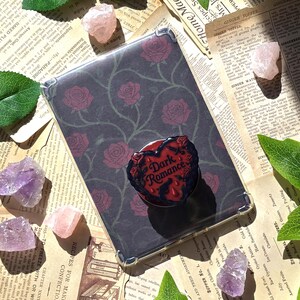 May include: A clear phone case with a dark background featuring a rose pattern. A red heart-shaped pop socket with the words "Dark Romance" is attached. Amethyst and rose quartz crystals are scattered around the case.