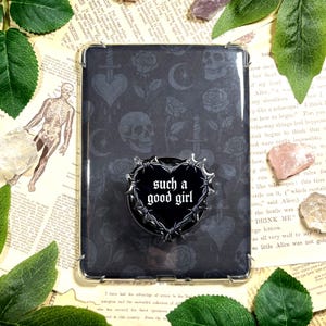May include: A black phone case with a repeating pattern of skulls, roses, hearts, and swords. A black heart-shaped phone grip with the text "such a good girl" is attached. The case is on a background of vintage book pages.