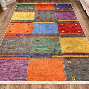 May include: A rectangular area rug with a patchwork design. The rug features squares in various colors, including orange, yellow, blue, red, and purple. Each square has small, square accents. The rug has a white border.