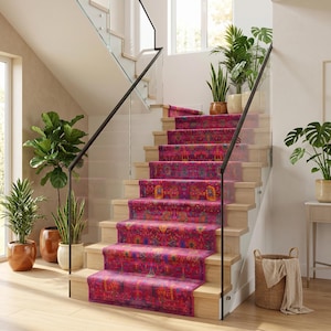 May include: A staircase with light wood steps features a vibrant magenta runner rug with a multi-colored floral and geometric pattern. The stairs have a glass railing and are surrounded by several potted green plants in a bright interior space.