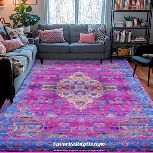 Può includere: Un tappeto a motivi rosa e blu con un design floreale. Il tappeto ha un centro giallo chiaro con un bordo blu più scuro. Il tappeto è in un soggiorno con un divano grigio e una libreria sullo sfondo.