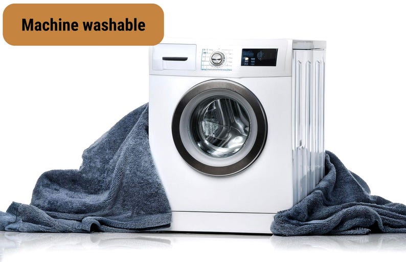 Puede incluir: Una lavadora blanca con una toalla azul drapeada sobre ella. La lavadora tiene una puerta plateada con una abertura redonda. El texto "Machine washable" es visible en la esquina superior izquierda de la imagen.