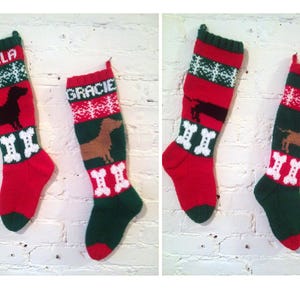 May include: Two Christmas stockings, one red and one green, with dog silhouettes and bone designs. One stocking says "BELLA" and the other says "GRACIE" in white letters. The stockings are hanging against a white brick wall.