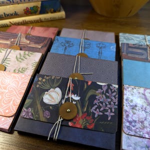 May include: A collection of rectangular paper folders featuring various patterns including floral, geometric, and vintage key designs. Each folder is secured with a string wrapped around a circular brown button fastener, creating a closure system.