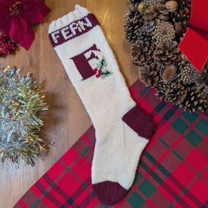 May include: Cream-colored knitted Christmas stocking with the name "FERN" in burgundy letters. The letter "F" is also in burgundy, with a holly sprig. The heel and toe are burgundy. The stocking is on a red plaid surface.