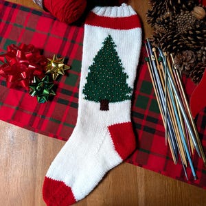 May include: A white knitted Christmas stocking featuring a green pine tree design with a brown trunk, accented by a red cuff, heel, and toe. The stocking is crafted with a ribbed top edge and a smooth knit body, showcasing a clear holiday-themed pattern.