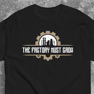 Può includere: T-shirt nero con grafica di ingranaggi e fabbrica in beige e bianco, e la scritta bianca "THE FACTORY MUST GROW". Il disegno è stampato sul davanti della maglietta.