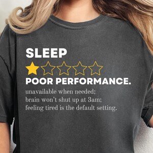 May include: Dark gray t-shirt with the text "SLEEP POOR PERFORMANCE." and a rating of one gold star and four empty stars. The text below reads "unavailable when needed; brain won't shut up at 3am; feeling tired is the default setting."