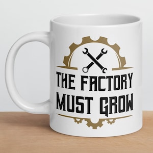 Peut inclure: Mug en céramique blanche avec l'inscription "THE FACTORY MUST GROW" en noir, ornée d'un engrenage et d'une clé à molette bruns. Le mug est posé sur une surface en bois.