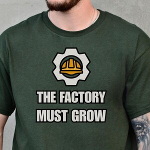 May include: Forest green t-shirt with the text "THE FACTORY MUST GROW" and a gear and hard hat graphic. A circular "FOREST GREEN" logo is at the bottom. The shirt is a short-sleeved crew neck.