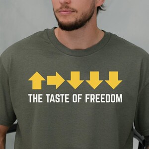 Può includere: T-shirt verde oliva con la scritta "THE TASTE OF FREEDOM" e grafiche di frecce gialle. La maglietta è di colore verde militare ed è in stile casual.