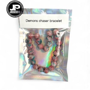 May include: A bracelet of pink, blue, and white speckled beads, shaped like a heart, is presented in a holographic bag. The bag has a white label with the text "Demons chaser bracelet". The bag is sealed.