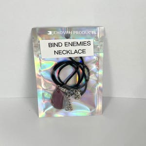 May include: A necklace with a dark purple teardrop-shaped pendant and a silver-colored charm, strung on a black cord. The necklace is packaged in a clear, iridescent pouch with the text "BIND ENEMIES NECKLACE". The pouch also has the text "JEHOVAH PRODUCTS".