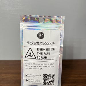 Może przedstawiać: Srebrna folia z etykietą w kolorze białym, na której widnieje napis "JEHOVAH PRODUCTS (A Subsidiary of FIHAJ LTD)" oraz "ENEMIES ON THE RUN SCRUB (Powerful effective scrub)". Etykieta zawiera również symbol ostrzegawczy, instrukcje użytkowania, adres strony internetowej, adres e-mail i trzy numery telefonów.