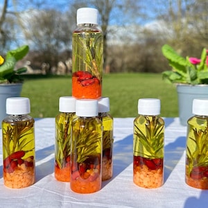 May include: Eight small clear glass bottles with white lids filled with a yellow liquid, red and orange pieces, and green sprigs. The bottles are arranged in a pyramid shape on a white surface.