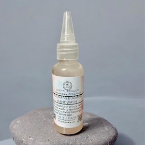 May include: A clear plastic bottle of handwash with a white label. The label reads "BUSINESS BOOMING HANDWASH" and includes the text "JEHOVAH PRODUCTS". The bottle has a clear, pointed applicator top. The bottle is sitting on a gray stone.