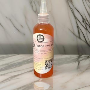 以下が含まれることがあります： 桃色の液体が詰まった透明なガラス瓶。瓶には、「STOP EVIL OIL」という黒い文字と「FIHAJ」という小さなロゴが書かれた白いラベルが貼られています。