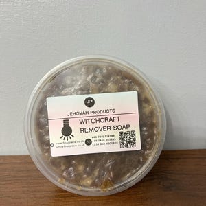 Pode incluir: Um recipiente de plástico transparente com uma barra de sabão marrom dentro. O rótulo diz "JEHOVAH PRODUCTS WITCHCRAFT REMOVER SOAP" com informações de contato e um código QR.