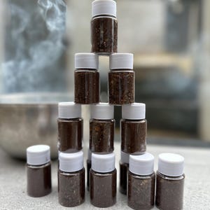 May include: A pyramid of small, clear glass jars with white lids, filled with a dark brown substance. The jars are arranged on a light gray surface, with smoke in the background. The jars are filled with a dark brown substance.