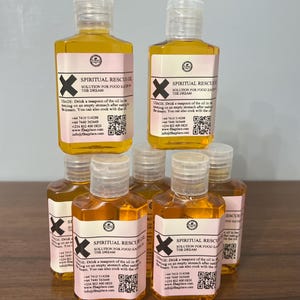 May include: A stack of clear plastic bottles filled with a golden liquid. Each bottle has a pink label with black text that reads "SPIRITUAL RESCUE OIL" and an "X" graphic. The bottles are arranged on a wooden surface.