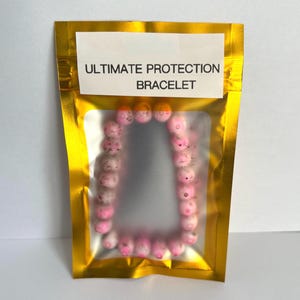 Pode incluir: Uma pulseira de contas rosa e brancas é exibida dentro de uma bolsa dourada com uma janela transparente. O texto "ULTIMATE PROTECTION BRACELET" é impresso em uma etiqueta branca acima da pulseira. As contas têm um design salpicado.