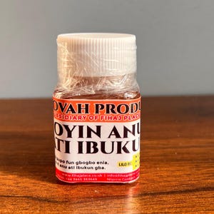May include: A small, clear glass bottle with a white cap, wrapped in clear plastic. The label features the text "OVAH PROD" and "OYIN AN TI IBUKU" in bold, white letters. The bottle contains a dark brown liquid. The background is a wooden surface.