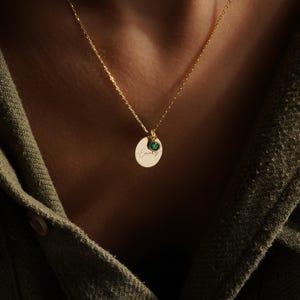 May include: A gold-colored necklace with a delicate chain. The necklace features a small, oval pendant with the name "Emily" engraved in cursive script. A small, round, green gemstone is set above the pendant, adding a pop of color.