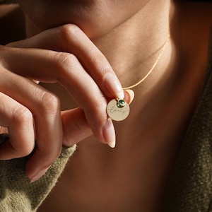 May include: Gold necklace with a round pendant engraved with the name "Emily" and a small green gemstone.