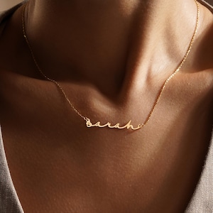 May include: A delicate gold necklace spelling out the name "Sarah" in a cursive script. The necklace is suspended from a fine chain, creating a minimalist and elegant look. The jewelry is worn around the neck.