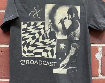 Puede incluir: Una camiseta negra con un diseño gráfico de estilo vintage que presenta a una persona cantando en un micrófono, un patrón de tablero de ajedrez, una estrella, un pie y la palabra "BROADCAST".