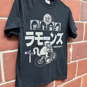 May include: Black t-shirt featuring a graphic design with the band members' portraits, Japanese characters, and a cat riding a motorcycle. The word "Ramones" is also printed on the shirt.