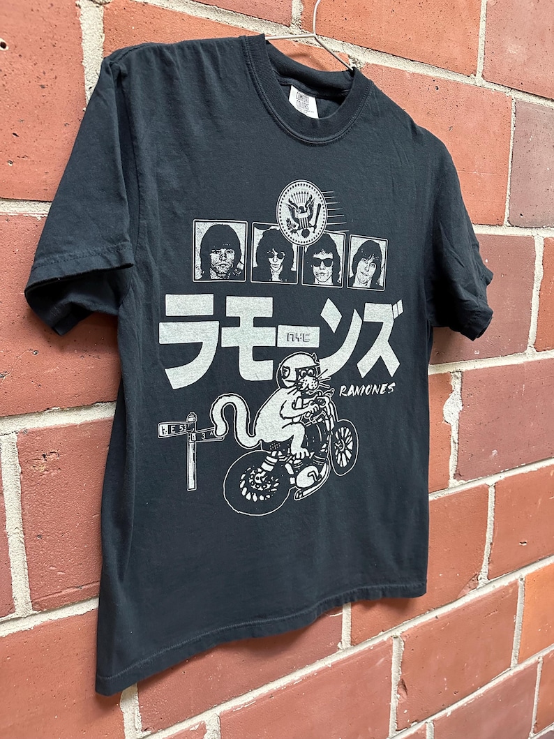 May include: Black t-shirt featuring a graphic design with the band members' portraits, Japanese characters, and a cat riding a motorcycle. The word "Ramones" is also printed on the shirt.