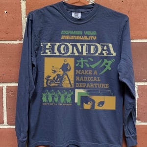 May include: Navy blue long-sleeve shirt with a graphic design featuring the word "HONDA" in a vintage font, a motorcycle illustration, and Japanese text. The shirt also has the phrases "Express Your Individuality" and "Make a Radical Departure."