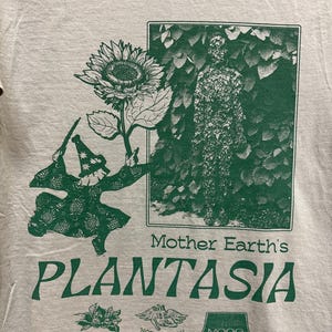 May include: A light gray t-shirt with a green graphic. The graphic includes a sunflower, a figure in a hat, and the text "Mother Earth's PLANTASIA". Below the text are additional small graphics and text.