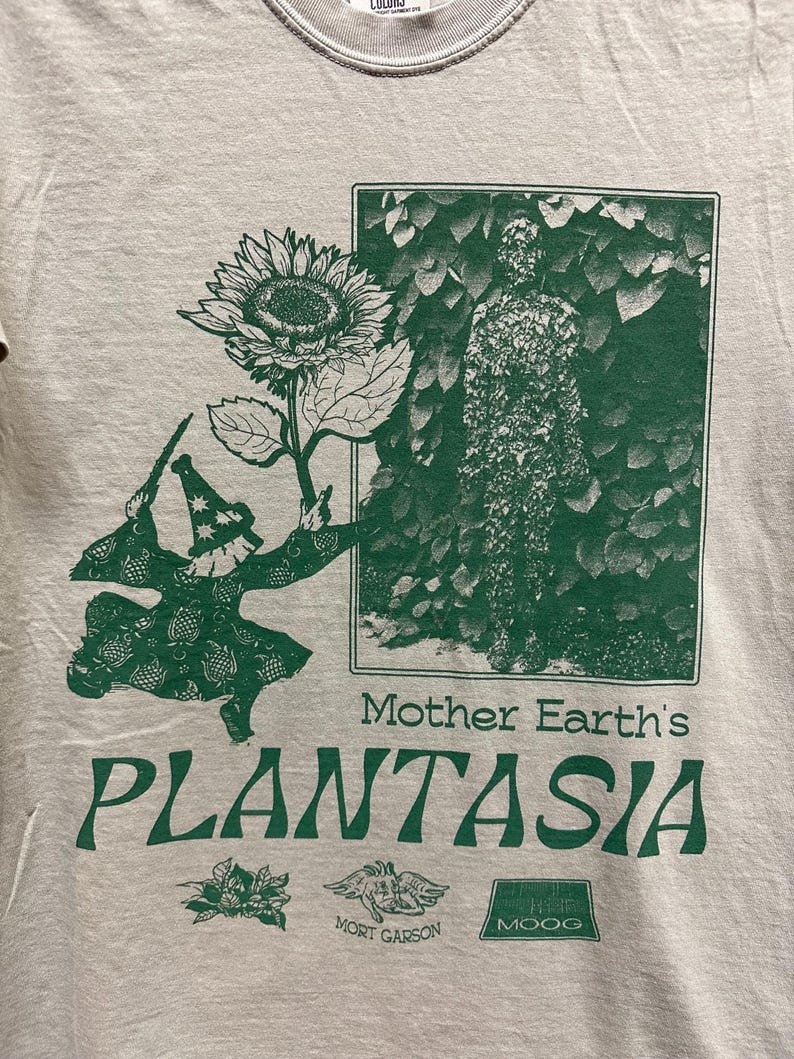 May include: A light gray t-shirt with a green graphic. The graphic includes a sunflower, a figure in a hat, and the text "Mother Earth's PLANTASIA". Below the text are additional small graphics and text.