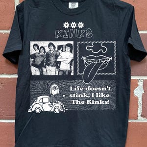 May include: A black short-sleeve t-shirt featuring a white graphic print. The design includes the text THE KINKS, a black and white photograph of the band, a line drawing of a mouth with a tongue sticking out, and a cartoon of a person on a car.
