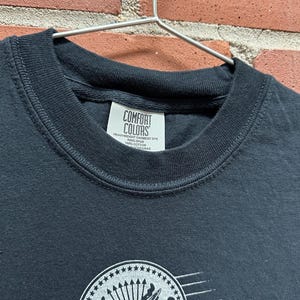 May include: A black Comfort Colors t-shirt with a crew neck. The shirt features a white graphic of a shield with an eagle, stars, and a baseball bat. The shirt is hanging on a metal hanger against a brick wall.