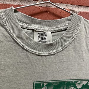 May include: Close-up of a gray Comfort Colors t-shirt hanging on a metal hanger. The shirt has a green graphic design on the front. The Comfort Colors label is visible at the neckline.