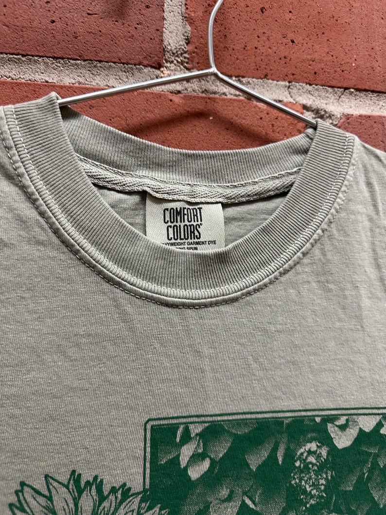 May include: Close-up of a gray Comfort Colors t-shirt hanging on a metal hanger. The shirt has a green graphic design on the front. The Comfort Colors label is visible at the neckline.