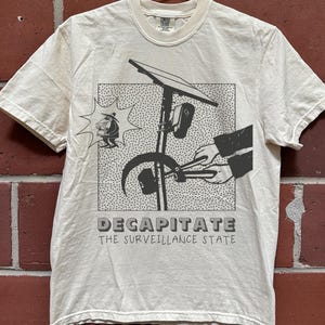 May include: A cream-colored short-sleeve t-shirt featuring a black graphic print of hands using bolt cutters on a surveillance camera pole. A small figure in a hat stands to the left. The text reads DECAPITATE THE SURVEILLANCE STATE.