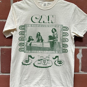 May include: A cream-colored short-sleeve t-shirt featuring a dark green graphic print. The design includes the text CAN above a framed photograph of a band, flanked by decorative borders, with a drum kit and musical note icons below.