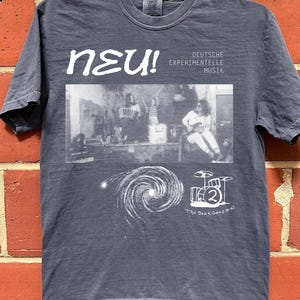 May include: A slate blue short-sleeve t-shirt featuring a white graphic print. The design includes the text "NEU! DEUTSCHE EXPERIMENTELLE MUSIK", a black and white photograph of a band, a spiral galaxy illustration, a drum kit, and "The Beat Goes on!"