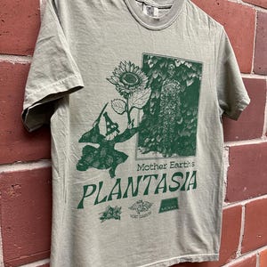 May include: Olive green t-shirt with a green graphic featuring a sunflower, a figure, and the text "Mother Earth's Plantasia." Additional text and logos are below the main text. The shirt is hanging on a metal hanger against a brick wall.