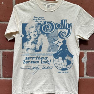 Può includere: Una t-shirt a maniche corte color crema con una stampa grafica blu. Il design include un ritratto di Dolly Parton, il testo "Dolly writes her own luck!", un'illustrazione di un globo, stelle e una firma. La maglietta ha un girocollo.