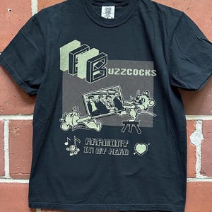 May include: Black t-shirt featuring a graphic design with the band name "Buzzcocks" in a block letter style. The design includes a cartoon mouse, a photo of the band, and the text "Harmony in my head".