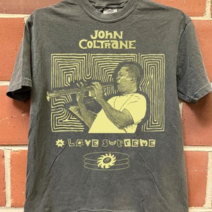 May include: A charcoal gray short-sleeve t-shirt featuring a light yellow graphic print of John Coltrane playing the saxophone. The design includes the text "JOHN COLTRANE" and "A LOVE SUPREME" with abstract line patterns and a sun icon.