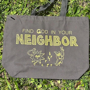 May include: A charcoal gray canvas tote bag featuring the yellow text FIND GOD IN YOUR NEIGHBOR with a graphic of a sun and a crescent moon character dancing with musical notes between them. The bag has two dark gray fabric shoulder straps.