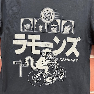 May include: Black t-shirt featuring a graphic of the Ramones band members, a cat riding a motorcycle, and Japanese text. The design includes the band's name and a NYC street sign. The shirt is made of a dark fabric.