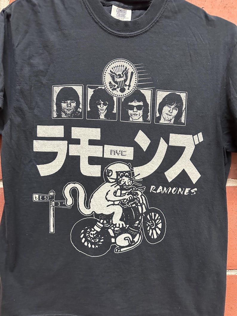 May include: Black t-shirt featuring a graphic of the Ramones band members, a cat riding a motorcycle, and Japanese text. The design includes the band's name and a NYC street sign. The shirt is made of a dark fabric.